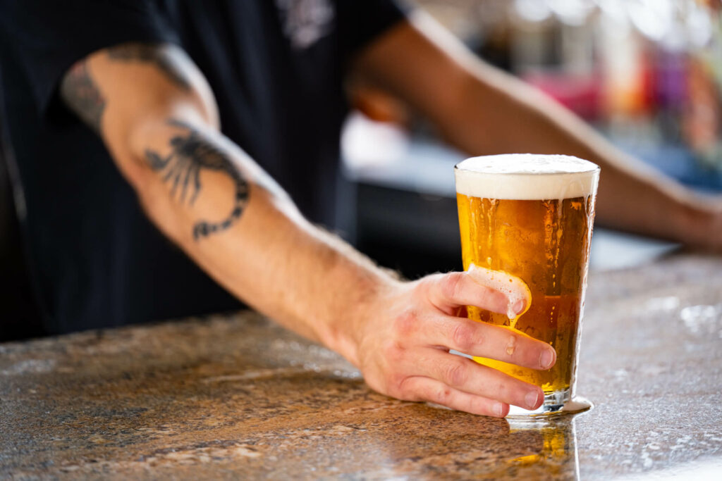 A person with a scorpion tattoo on their forearm is placing a glass of beer with a foamy head on a bar counter.