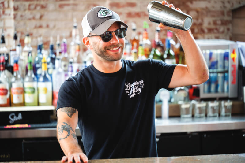 A smiling bartender wearing sunglasses, a baseball cap, and a 