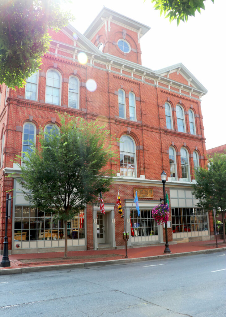 A historic red brick building with large arched windows, trees and hanging flower baskets lining the sidewalk, and several flags displayed above the entrance on a sunny day.
