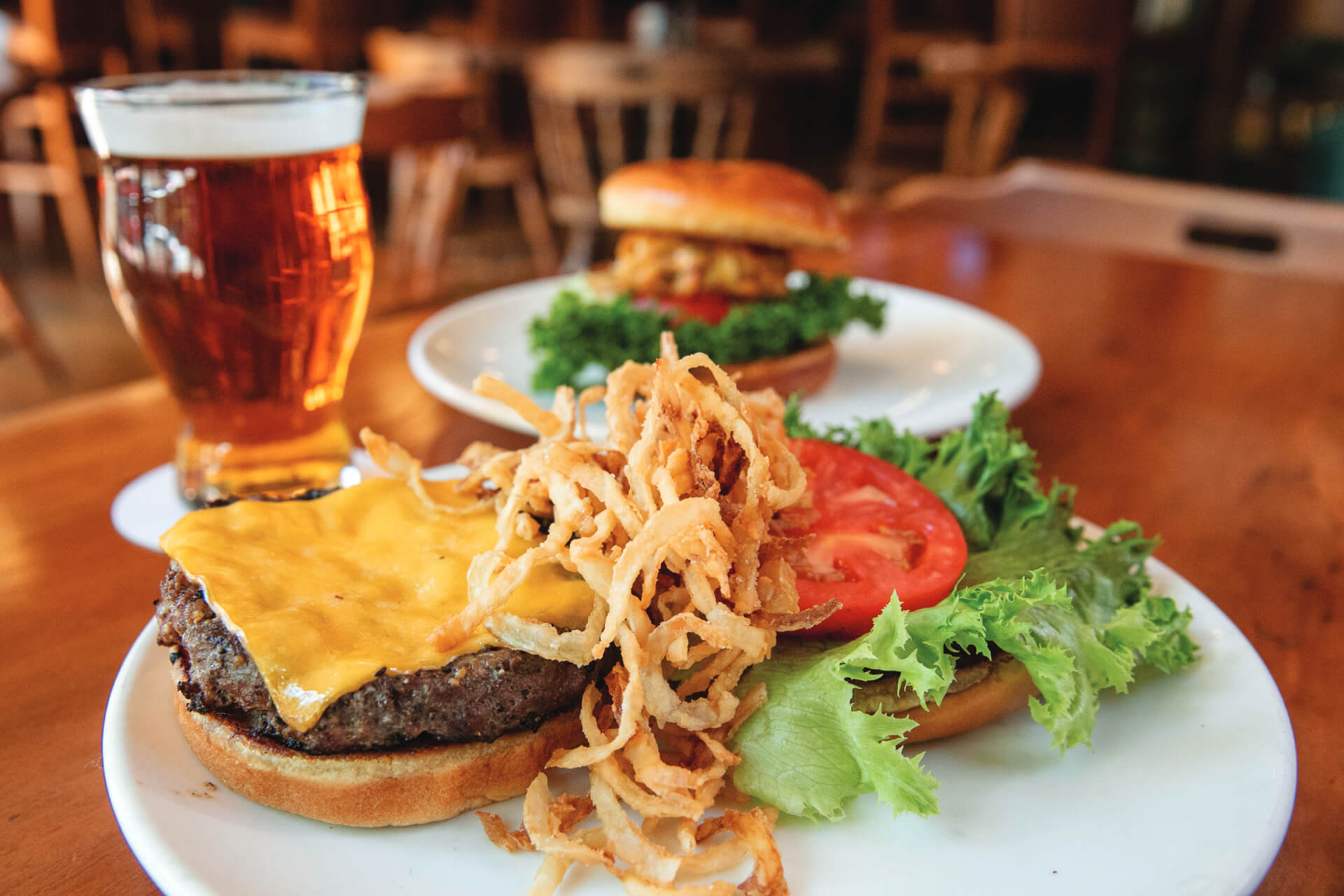 A close-up of a cheeseburger topped with crispy fried onions, lettuce, and tomato on a white plate, next to a glass of beer; another burger is blurred in the background.