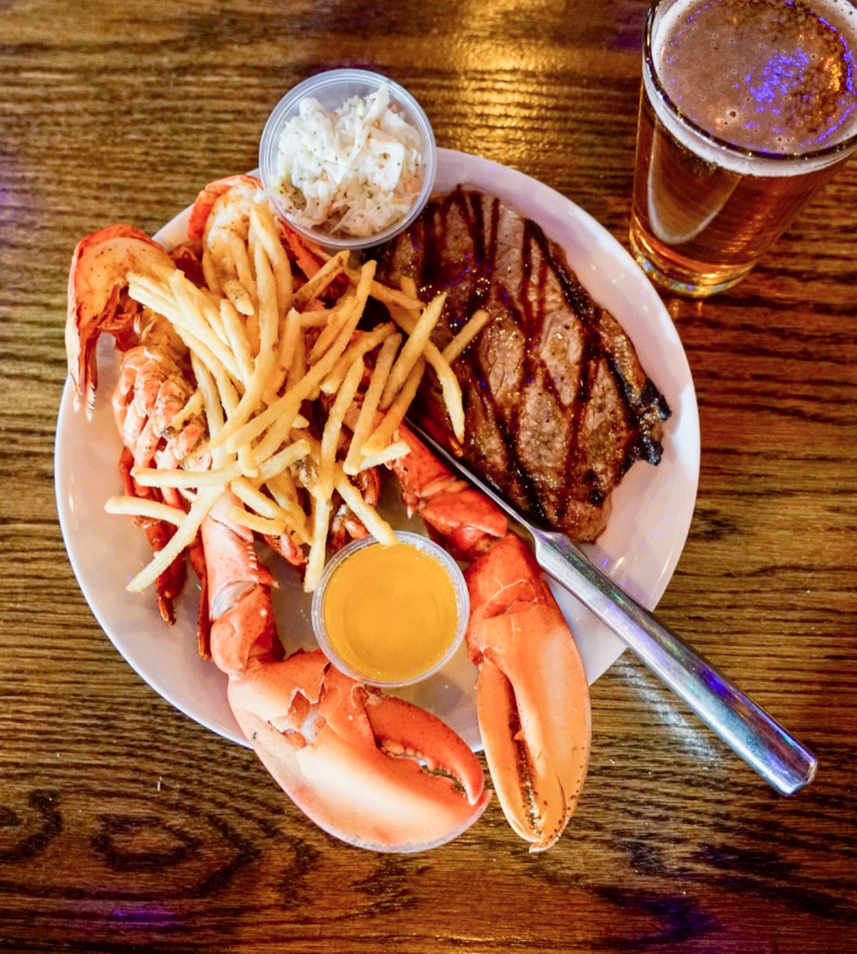A plate with grilled steak, lobster claws with a cup of butter, french fries, and a side of coleslaw. A glass of beer sits next to the plate on a wooden table.