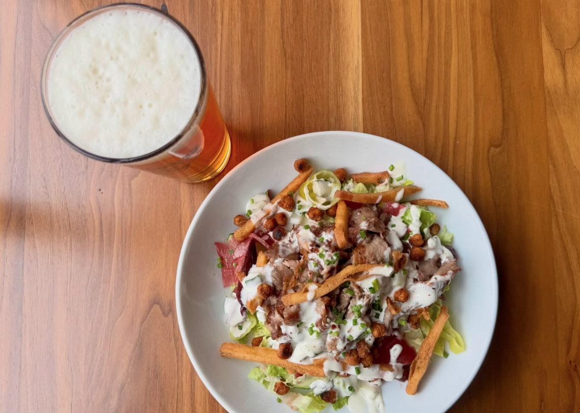 A glass of frothy beer sits next to a bowl of salad topped with grilled meat, crispy bread sticks, lettuce, chopped chives, and creamy dressing, served on a wooden table.