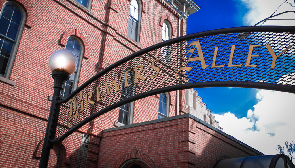 A curved metal archway with gold lettering reads 'Brewer's Alley' in front of a red brick building on a sunny day, with a decorative lamp post and blue sky with clouds in the background.