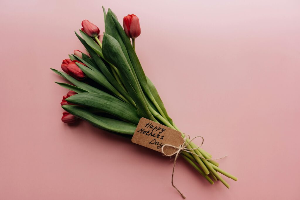 A bouquet of red tulips with green leaves lies on a pink surface. A brown tag tied with twine to the stems reads 'Happy Mother’s Day' in handwritten lettering.
