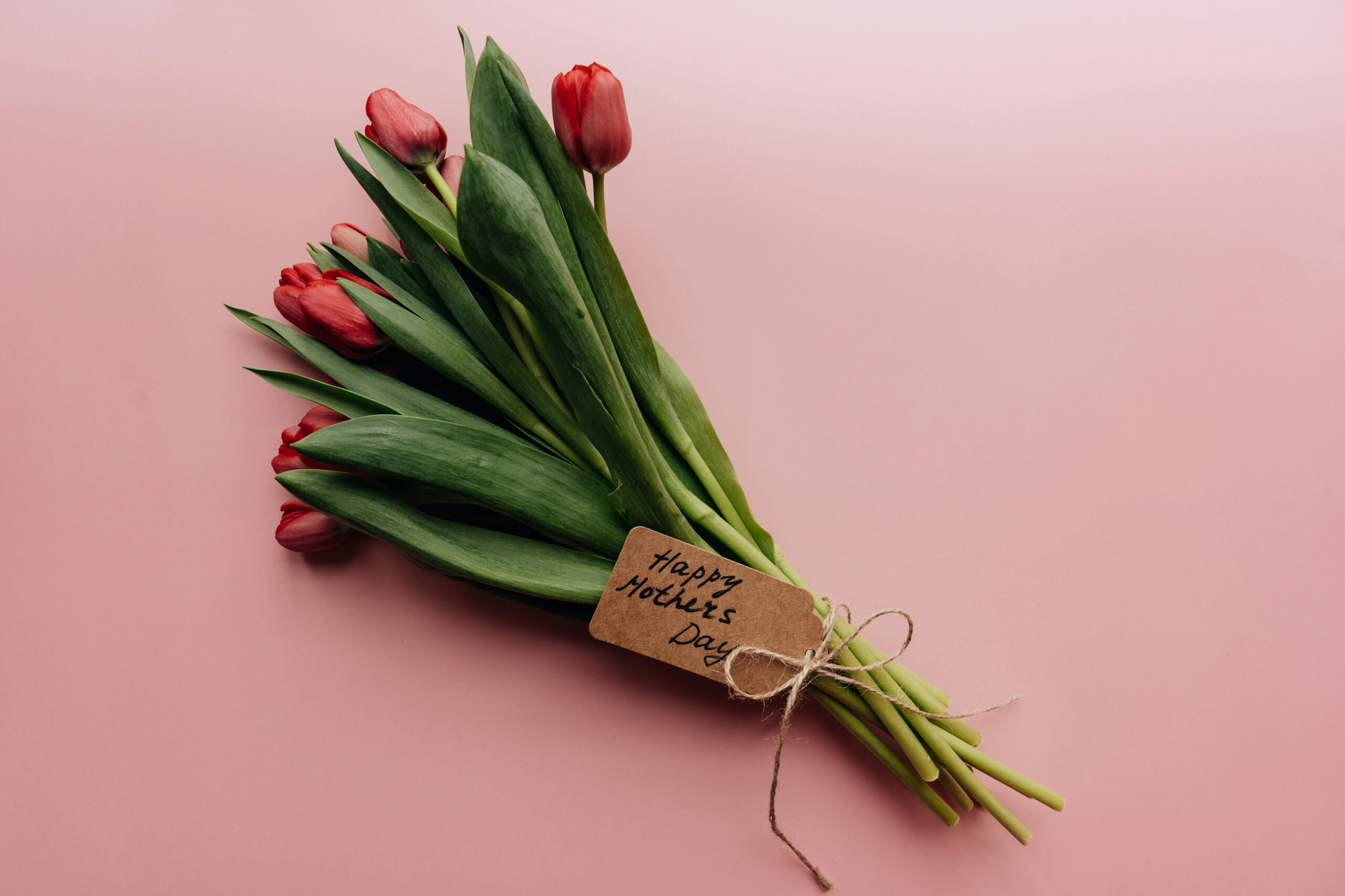 A bouquet of red tulips with green leaves lies on a pink surface. A brown tag tied with twine to the stems reads "Happy Mother’s Day" in handwritten lettering.