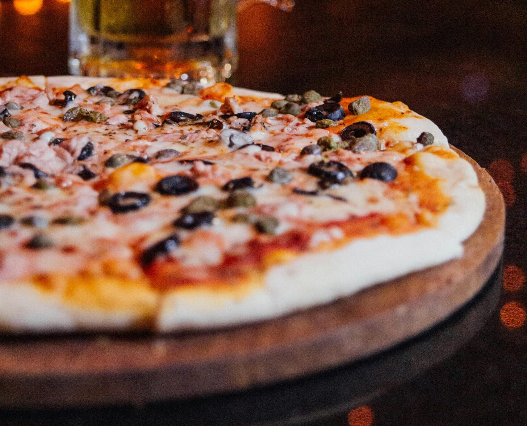 A close-up of a pizza topped with black olives, mushrooms, and cheese on a wooden board, with a frosty glass of beer in the background on a dimly lit table.