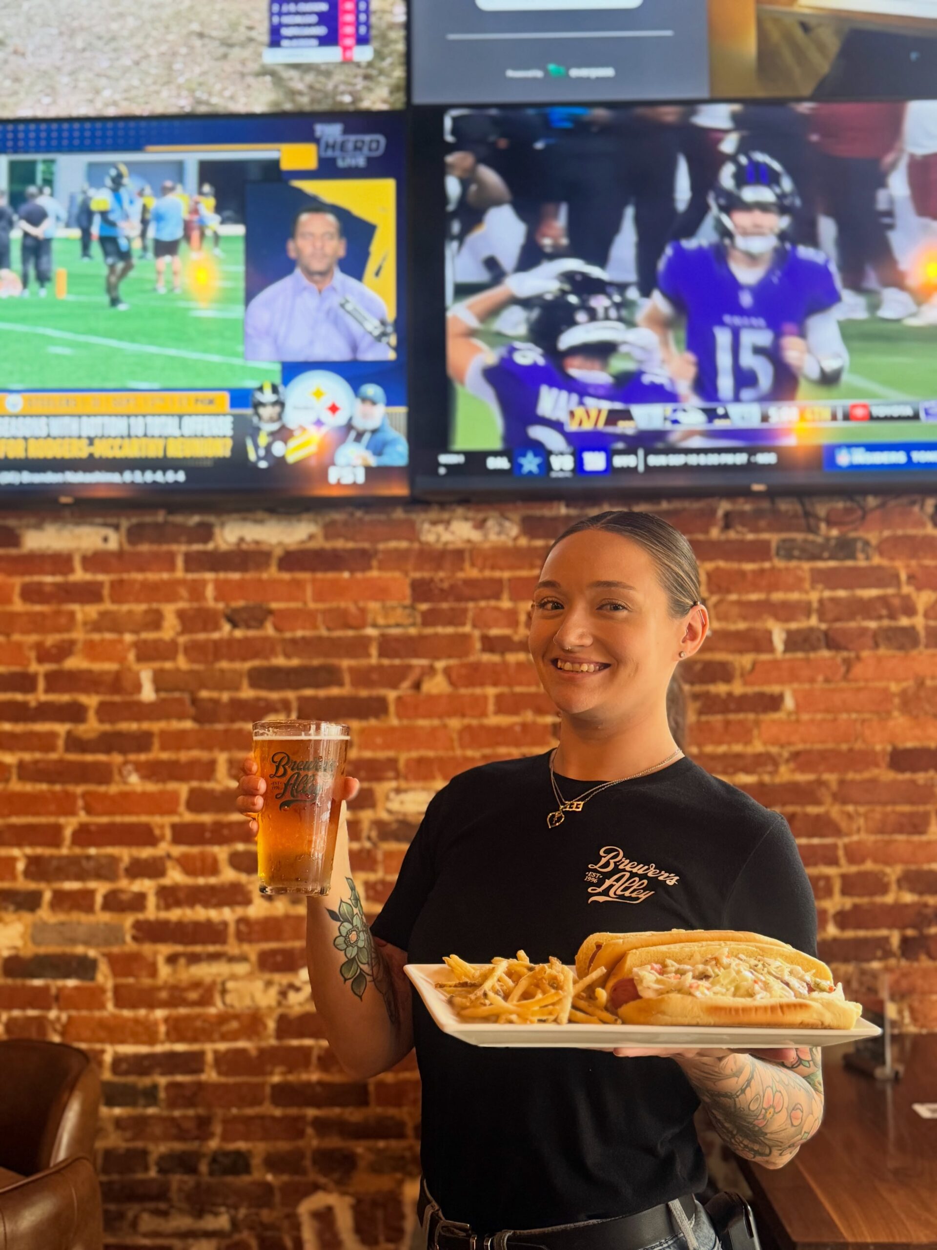 A smiling server holds a pint of beer and a plate with a sandwich and fries in front of two TV screens showing sports games, with a brick wall in the background.