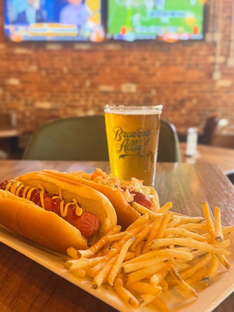 A plate with two hot dogs topped with mustard and relish, a pile of thin fries, and a glass of beer sits on a wooden table in a restaurant with brick walls and TV screens in the background.