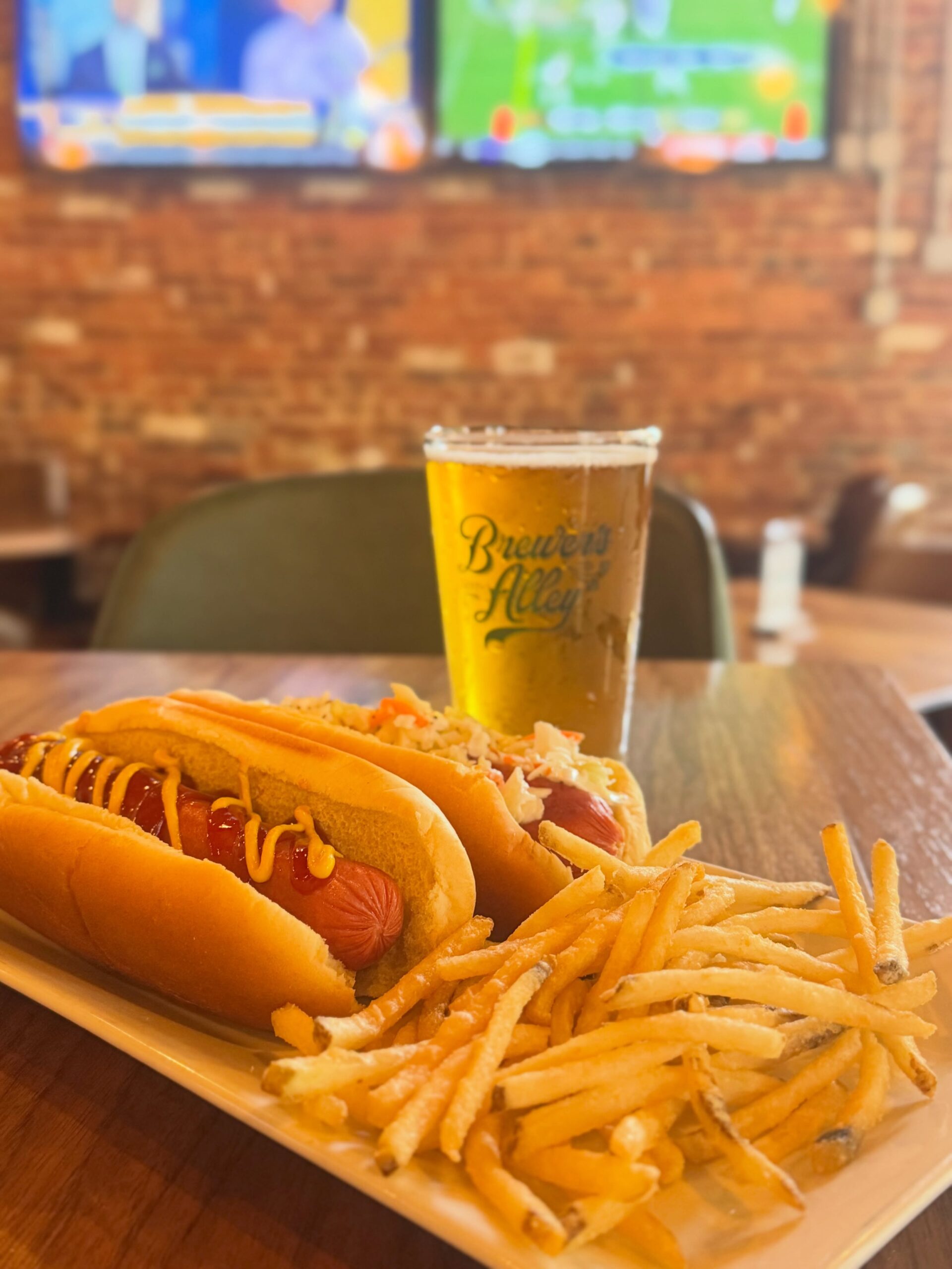 A plate with two hot dogs topped with mustard and relish, a pile of thin fries, and a glass of beer sits on a wooden table in a restaurant with brick walls and TV screens in the background.