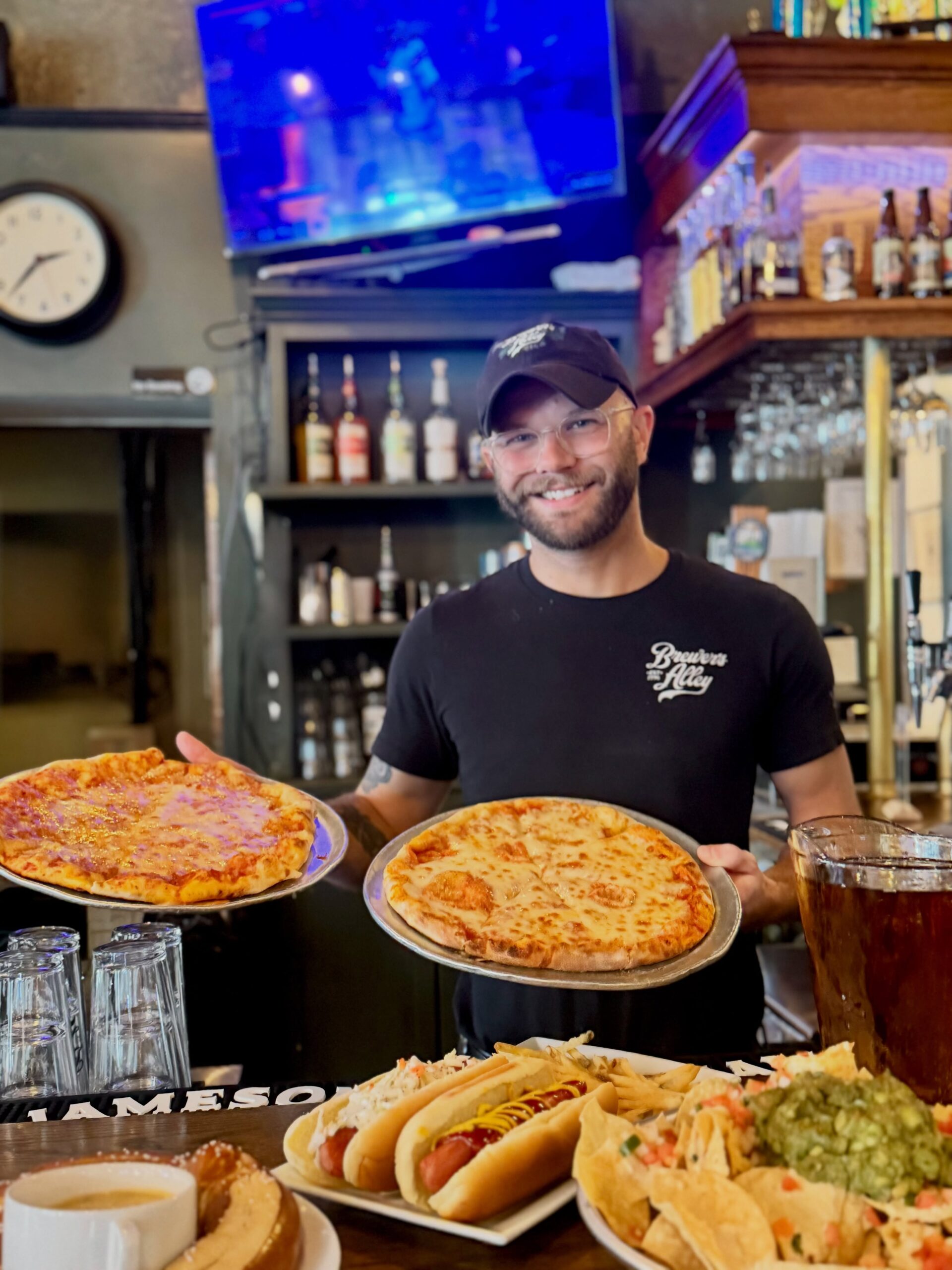 A smiling person wearing glasses and a cap holds two pizzas behind a bar, with tacos, guacamole, and drinks on the counter in front of them. Bottles and a TV are in the background.