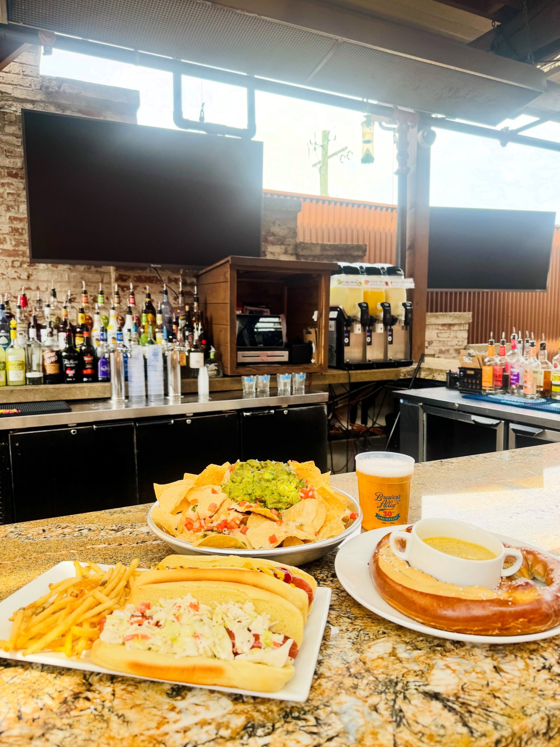 Three plates of food—nachos with guacamole, hot dogs with fries, a pretzel with cheese—sit on a bar counter with beer, liquor bottles, and empty TV screens in the background.